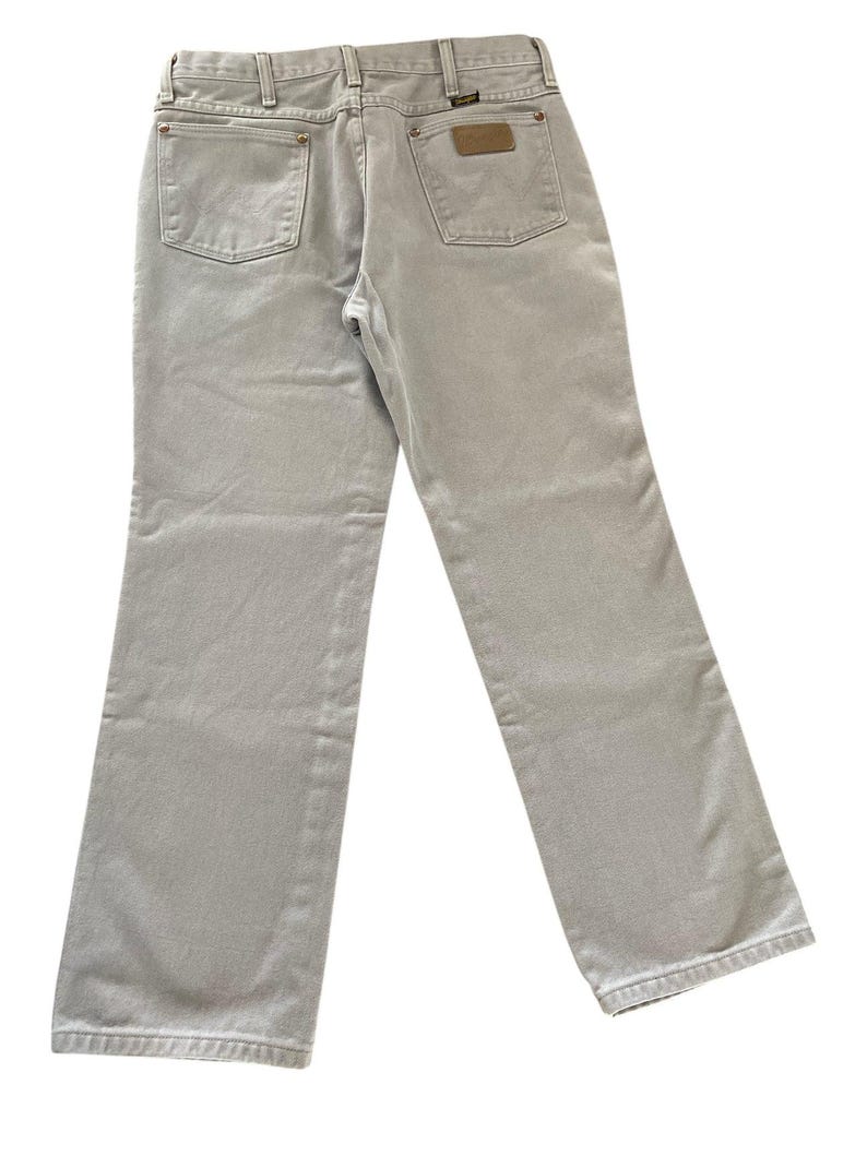 Op de afbeelding: Lichtgrijze denim jeans met een klassiek ontwerp met vijf zakken. De jeans hebben een bruine leren patch op de achterzak en een metalen knoopsluiting. Het merk Wrangler is zichtbaar.