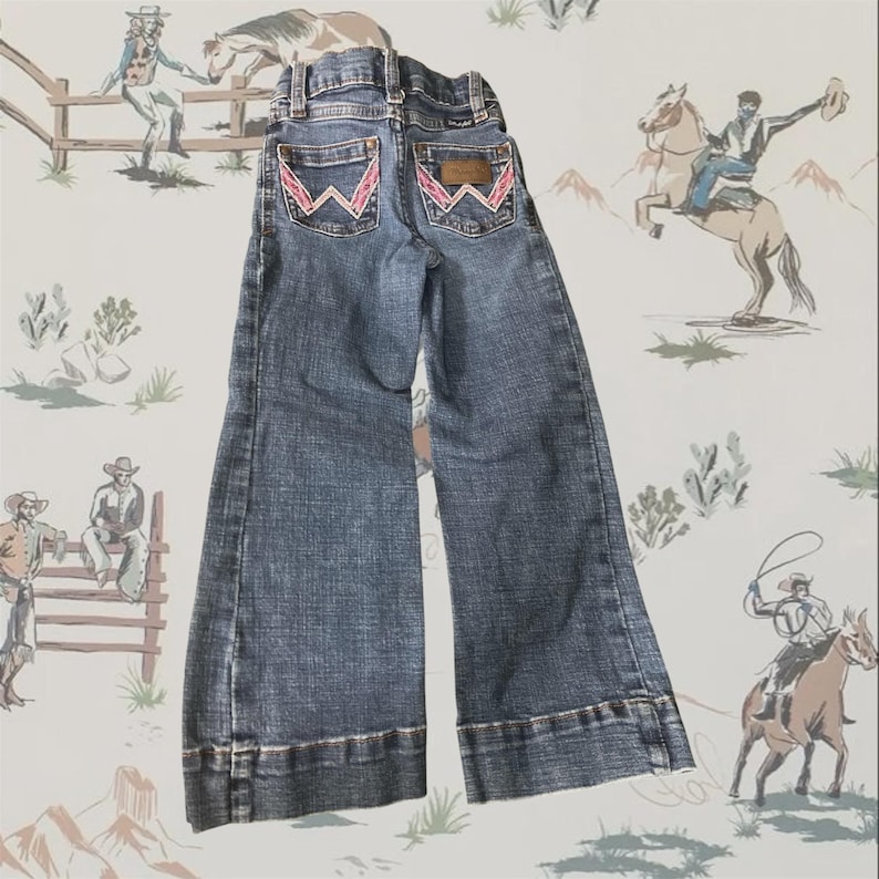 May include: Blue denim jeans featuring a wide-leg cut and a high-rise waist. The back pockets are detailed with a signature W-shaped embroidery in pink stitching. A brown leather patch is positioned above the right back pocket with Wrangler branding.