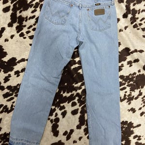 May include: Pale blue denim jeans featuring the Wrangler logo on the back pockets. These jeans have a classic five-pocket design and a straight leg fit. They are made from a durable denim material, suitable for everyday wear. The jeans are a light wash.
