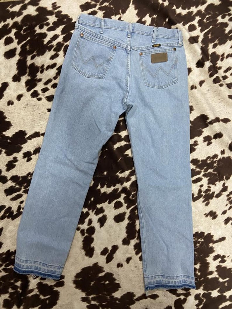 May include: Pale blue denim jeans featuring the Wrangler logo on the back pockets. These jeans have a classic five-pocket design and a straight leg fit. They are made from a durable denim material, suitable for everyday wear. The jeans are a light wash.