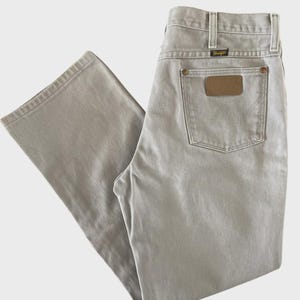 Op de afbeelding: Lichtgrijze Wrangler-jeans met een klassiek ontwerp met vijf zakken. De jeans hebben een bruine leren patch op de achterzak en een klein zwart label boven de achterzak. De jeans zijn gemaakt van een duurzame stof.