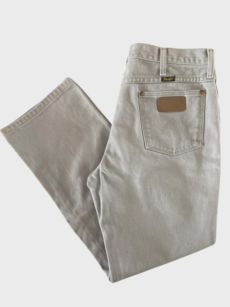 Op de afbeelding: Lichtgrijze Wrangler-jeans met een klassiek ontwerp met vijf zakken. De jeans hebben een bruine leren patch op de achterzak en een klein zwart label boven de achterzak. De jeans zijn gemaakt van een duurzame stof.