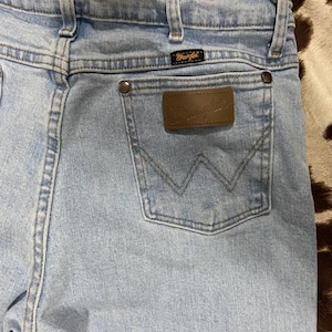 May include: Light blue denim jeans with a brown leather patch and the Wrangler logo. The jeans have a classic five-pocket design, belt loops, and a back pocket with a stitched 'W' design. The jeans are made of a durable denim material.