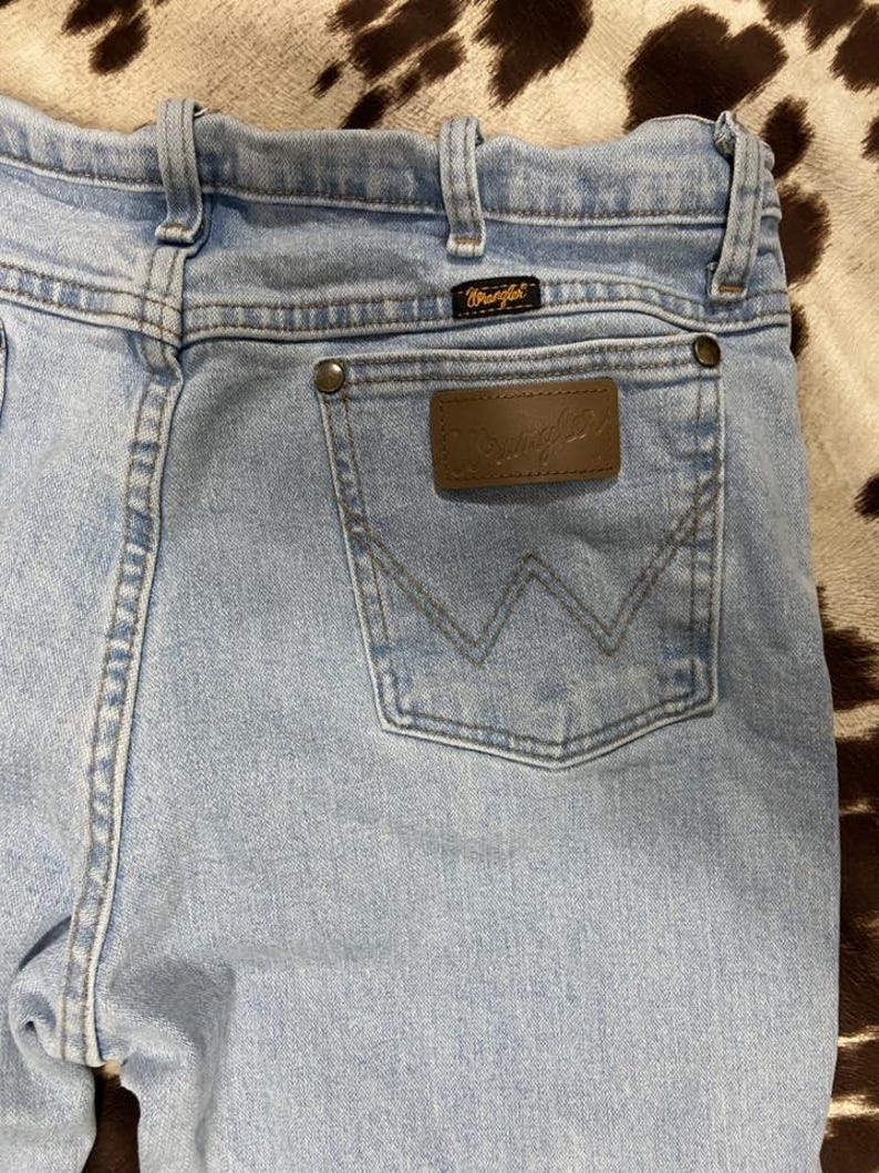May include: Light blue denim jeans with a brown leather patch and the Wrangler logo. The jeans have a classic five-pocket design, belt loops, and a back pocket with a stitched 'W' design. The jeans are made of a durable denim material.