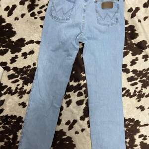 May include: Light blue denim jeans with the Wrangler logo on the back pocket. The jeans have a classic five-pocket design and a straight leg cut. The jeans are laid flat on a brown and white patterned surface.