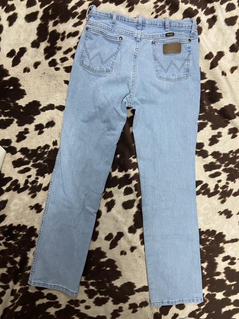 May include: Light blue denim jeans with the Wrangler logo on the back pocket. The jeans have a classic five-pocket design and a straight leg cut. The jeans are laid flat on a brown and white patterned surface.