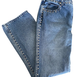 May include: Light blue denim jeans with silver-colored stud accents along the front pockets, side seams, and waistband. The jeans have a classic five-pocket design and a straight leg cut. The denim has a faded wash.