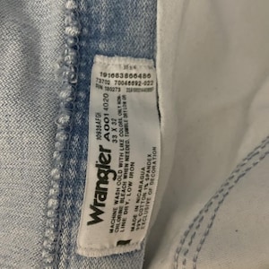 May include: Close-up of light blue Wrangler jeans. The jeans have a white tag with the Wrangler logo and care instructions. The tag also includes the size 33 x 32. The jeans are made of 99% cotton and 1% spandex.
