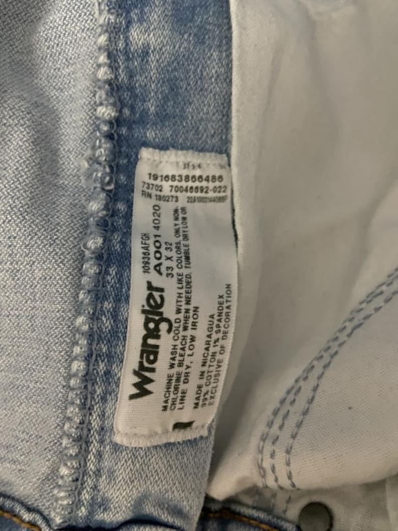 May include: Close-up of light blue Wrangler jeans. The jeans have a white tag with the Wrangler logo and care instructions. The tag also includes the size 33 x 32. The jeans are made of 99% cotton and 1% spandex.