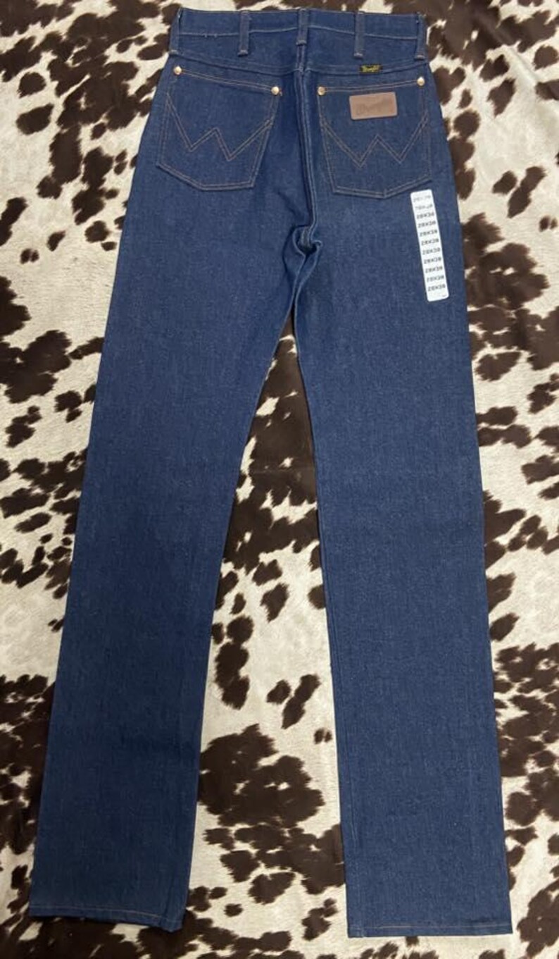 May include: Dark blue denim jeans with the Wrangler logo on the back pockets and a leather patch. The jeans have a classic five-pocket design and a straight leg. The tag shows the size.