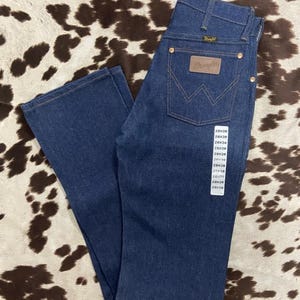 May include: Dark blue denim Wrangler jeans with a brown leather patch and the Wrangler logo on the back pocket. The jeans have a classic five-pocket design and a straight leg. The size tag shows a 28x38 size.
