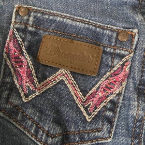 May include: Blue denim jeans featuring a back pocket with a brown leather Wrangler patch and a distinctive W-shaped embroidery pattern in pink and white thread. The pocket corners are secured with copper-colored metal rivets featuring a W logo.
