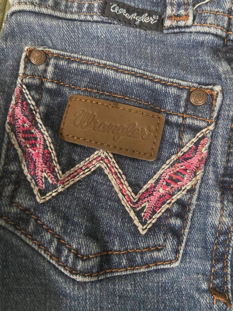 May include: Blue denim jeans featuring a back pocket with a brown leather Wrangler patch and a distinctive W-shaped embroidery pattern in pink and white thread. The pocket corners are secured with copper-colored metal rivets featuring a W logo.