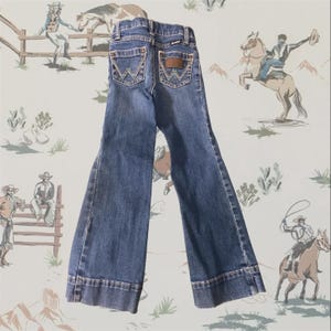 Op de afbeelding: Een donkerblauwe spijkerbroek van denim met uitlopende broekspijpen. De achterkant van de broek heeft twee zakken met een kenmerkend geel stiksel in de vorm van een W en een bruine leren patch op de rechterzak met de merknaam Wrangler.