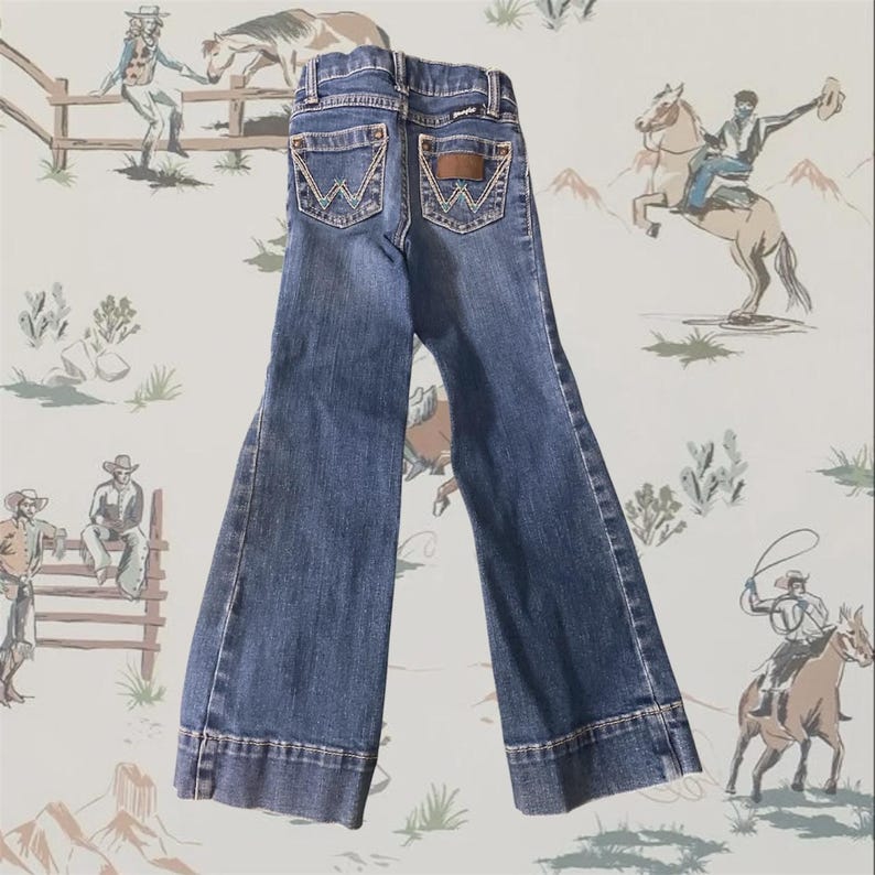 Op de afbeelding: Een donkerblauwe spijkerbroek van denim met uitlopende broekspijpen. De achterkant van de broek heeft twee zakken met een kenmerkend geel stiksel in de vorm van een W en een bruine leren patch op de rechterzak met de merknaam Wrangler.