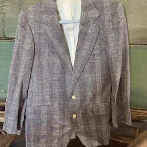 May include: A tweed blazer with a plaid pattern in shades of brown, gray, and burgundy hangs on a hanger. The jacket has two buttons and two front pockets. The interior lining is a cream color.