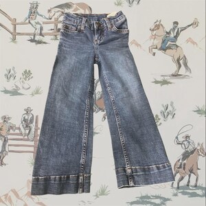 May include: A pair of medium-wash blue denim jeans featuring a wide-leg silhouette, a classic five-pocket design, a button and zipper fly closure, and visible copper-toned stitching along the seams and the wide hem at the bottom of the legs.