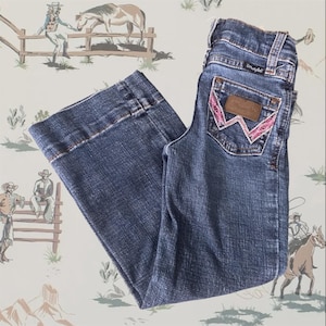 May include: Medium-wash blue denim jeans featuring a wide-leg cut and a high-rise waist. The back pocket displays a brown leather patch with the Wrangler logo and a distinctive W-shaped embroidery stitched in bright pink thread.