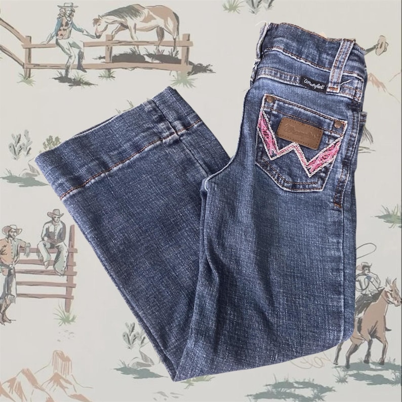 May include: Medium-wash blue denim jeans featuring a wide-leg cut and a high-rise waist. The back pocket displays a brown leather patch with the Wrangler logo and a distinctive W-shaped embroidery stitched in bright pink thread.