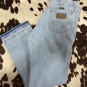 May include: Pale blue denim jeans with a classic five-pocket design. Features a brown leather patch on the back pocket and the Wrangler logo. The jeans have a straight leg cut and a faded wash, giving them a vintage look.