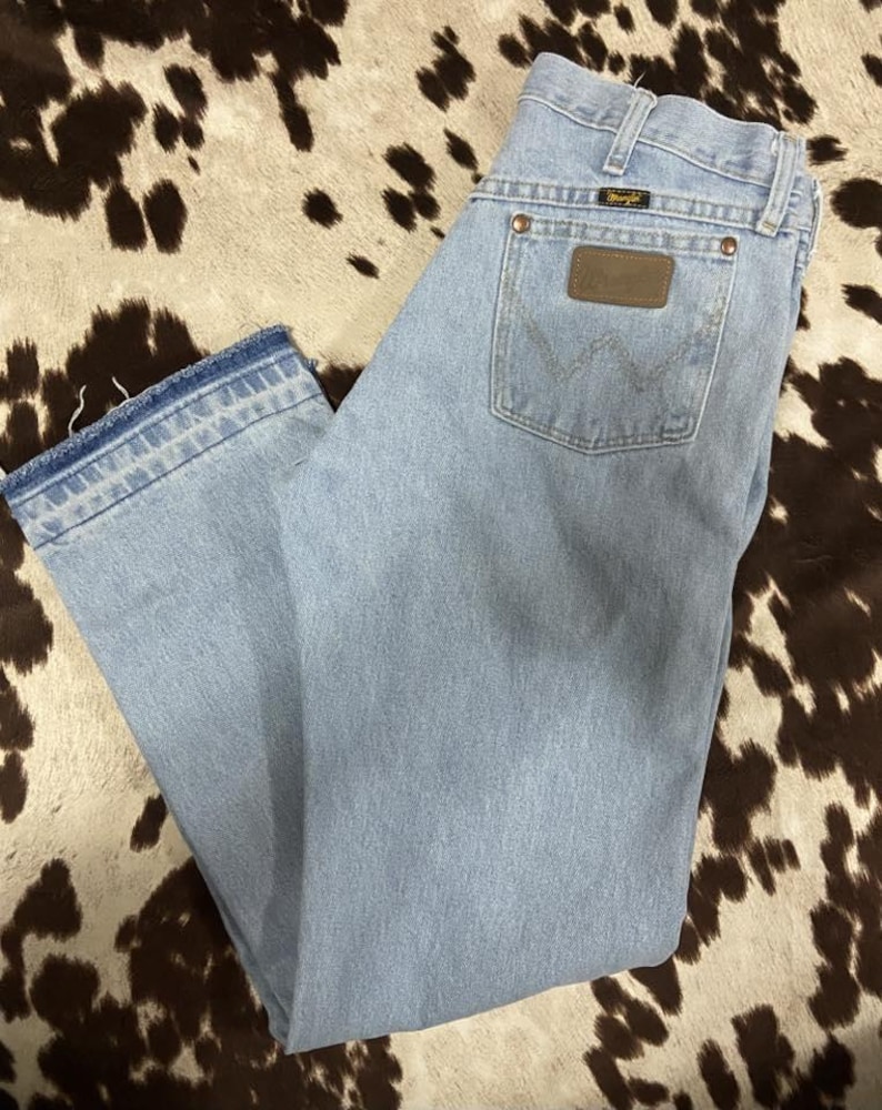 May include: Pale blue denim jeans with a classic five-pocket design. Features a brown leather patch on the back pocket and the Wrangler logo. The jeans have a straight leg cut and a faded wash, giving them a vintage look.