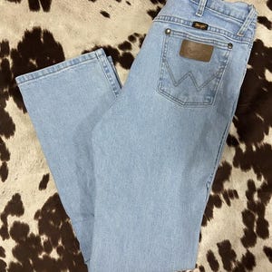 May include: Light blue denim jeans with a classic five-pocket design. The jeans feature a brown leather patch on the back pocket with the brand name. The jeans are laid flat, showcasing the light wash and straight leg cut.