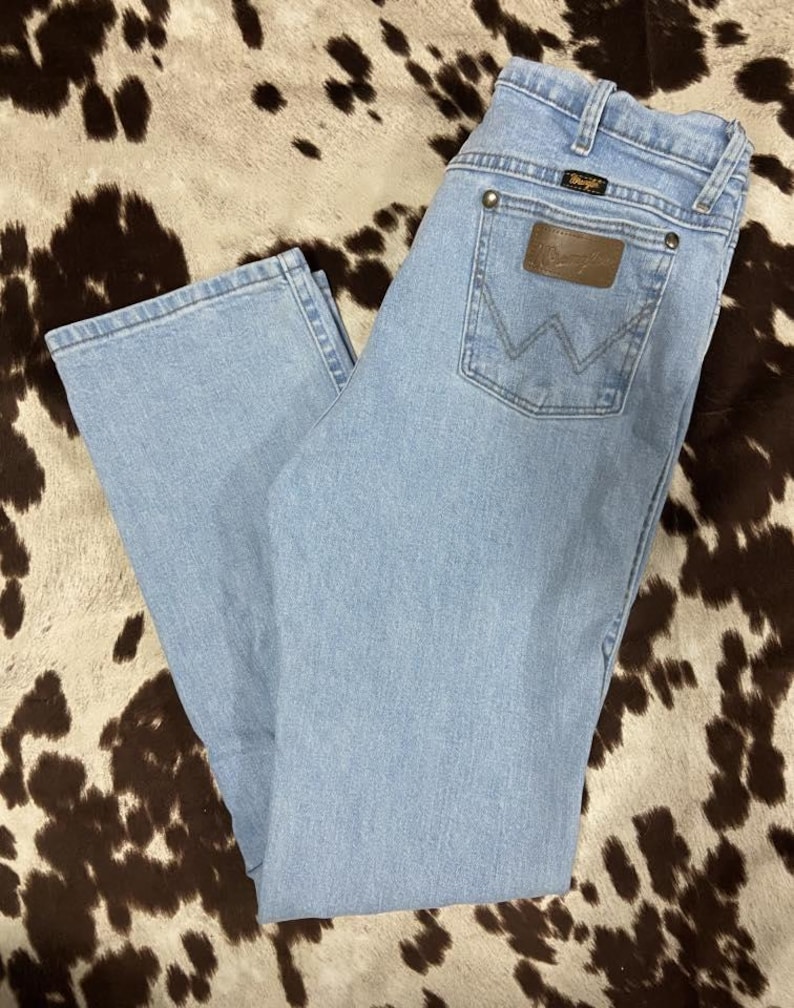 May include: Light blue denim jeans with a classic five-pocket design. The jeans feature a brown leather patch on the back pocket with the brand name. The jeans are laid flat, showcasing the light wash and straight leg cut.