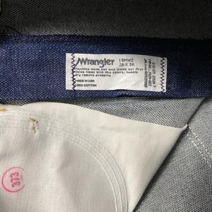 May include: Close-up of a pair of blue denim jeans. The Wrangler label is visible, with text indicating the style, size 28 x 36, and care instructions. The jeans are made of 100% cotton and made in the USA.