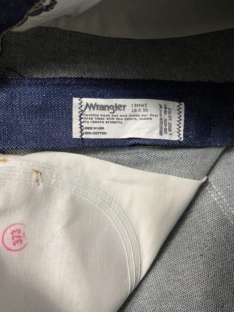 May include: Close-up of a pair of blue denim jeans. The Wrangler label is visible, with text indicating the style, size 28 x 36, and care instructions. The jeans are made of 100% cotton and made in the USA.