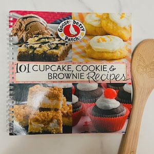 May include: A spiral-bound cookbook titled 101 Cupcake, Cookie & Brownie Recipes by Gooseberry Patch, featuring photographs of baked goods on the cover, placed next to a light-colored wooden spoon with a butterfly engraving on the handle.