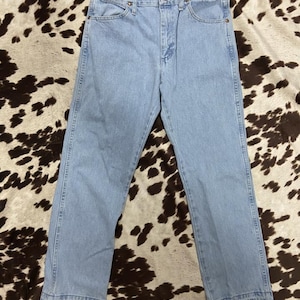 May include: Pale blue denim jeans with a traditional five-pocket design. The jeans feature a straight leg cut and a button and zip fastening. The hemline is frayed. The jeans are shown on a brown and white patterned surface.