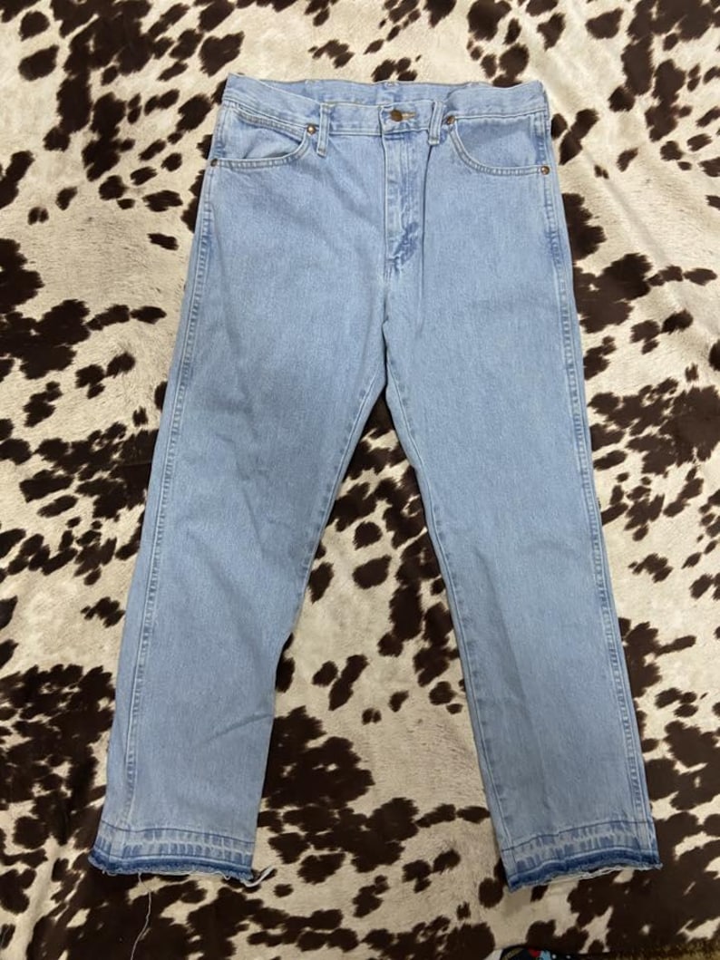 May include: Pale blue denim jeans with a traditional five-pocket design. The jeans feature a straight leg cut and a button and zip fastening. The hemline is frayed. The jeans are shown on a brown and white patterned surface.
