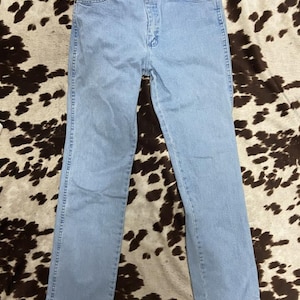 May include: Light blue denim jeans with a classic five-pocket design. The jeans have a straight leg cut and a button and zipper closure. The jeans are laid flat on a cowhide patterned surface.