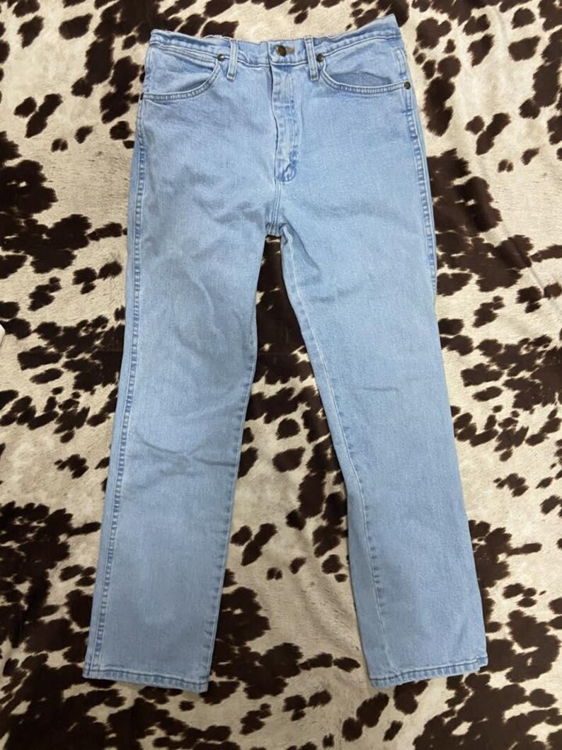 May include: Light blue denim jeans with a classic five-pocket design. The jeans have a straight leg cut and a button and zipper closure. The jeans are laid flat on a cowhide patterned surface.