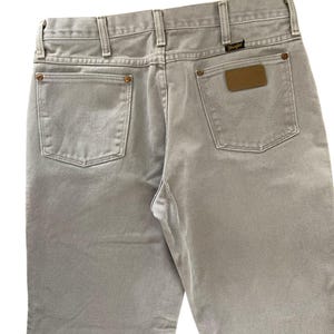 Op de afbeelding: Lichtgrijze Wrangler-jeans, gezien vanaf de achterkant. Voorzien van twee achterzakken, riemlussen en een bruin leren patch met het Wrangler-logo. De jeans hebben een klassieke, rechte pijp.