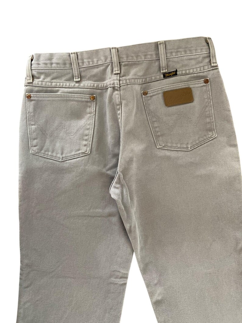 Op de afbeelding: Lichtgrijze Wrangler-jeans, gezien vanaf de achterkant. Voorzien van twee achterzakken, riemlussen en een bruin leren patch met het Wrangler-logo. De jeans hebben een klassieke, rechte pijp.