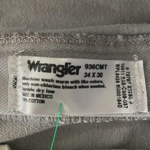 Op de afbeelding: Close-up van een lichtgrijs Wrangler jeanslabel. Het label toont de merknaam, maat 34 x 30 en wasvoorschriften. De jeans is gemaakt in Mexico en is 100% katoen.