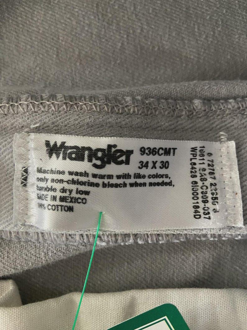 Op de afbeelding: Close-up van een lichtgrijs Wrangler jeanslabel. Het label toont de merknaam, maat 34 x 30 en wasvoorschriften. De jeans is gemaakt in Mexico en is 100% katoen.