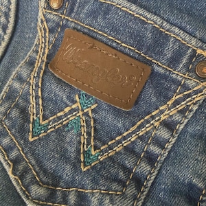 Op de afbeelding: Een blauwe spijkerbroekzak met een bruine leren patch waarop de tekst Wrangler in reli&euml;f staat. De zak is voorzien van beige contraststiksels en een decoratief W-vormig borduurpatroon in een levendige kleur blauwgroen.