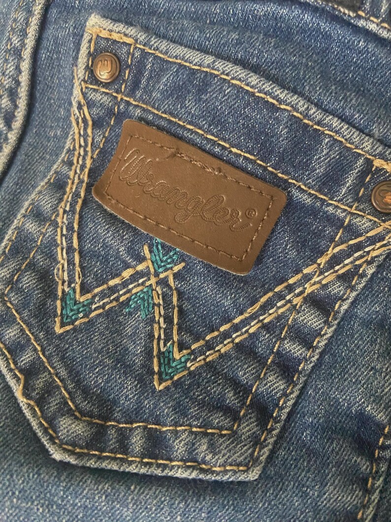 Op de afbeelding: Een blauwe spijkerbroekzak met een bruine leren patch waarop de tekst Wrangler in reli&euml;f staat. De zak is voorzien van beige contraststiksels en een decoratief W-vormig borduurpatroon in een levendige kleur blauwgroen.
