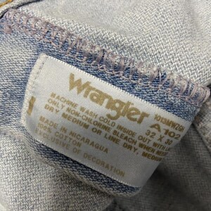 May include: Close-up of light blue denim jeans with a Wrangler label. The label displays the brand name, size 32 x 30, and washing instructions. The jeans are made of 100% cotton and made in Nicaragua.