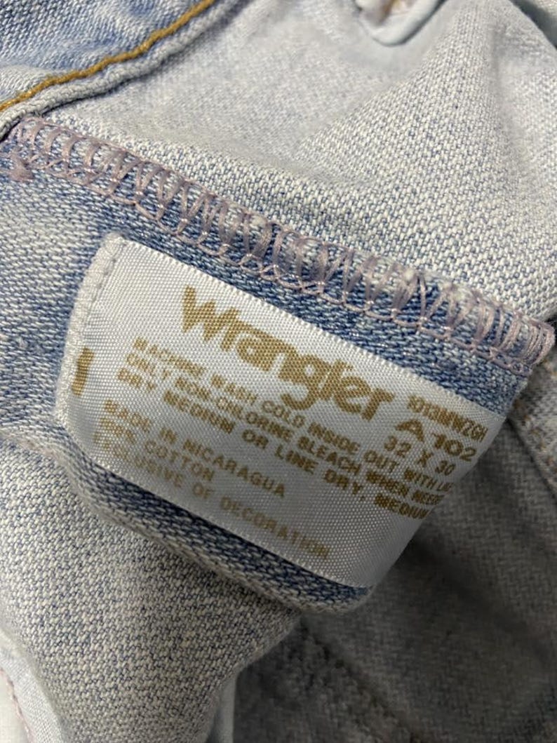 May include: Close-up of light blue denim jeans with a Wrangler label. The label displays the brand name, size 32 x 30, and washing instructions. The jeans are made of 100% cotton and made in Nicaragua.