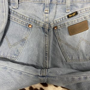 May include: Close-up of light blue denim jeans. The jeans feature classic five-pocket styling, copper rivets, and a brown leather Wrangler patch. The Wrangler label is visible on the back pocket. The jeans are folded.