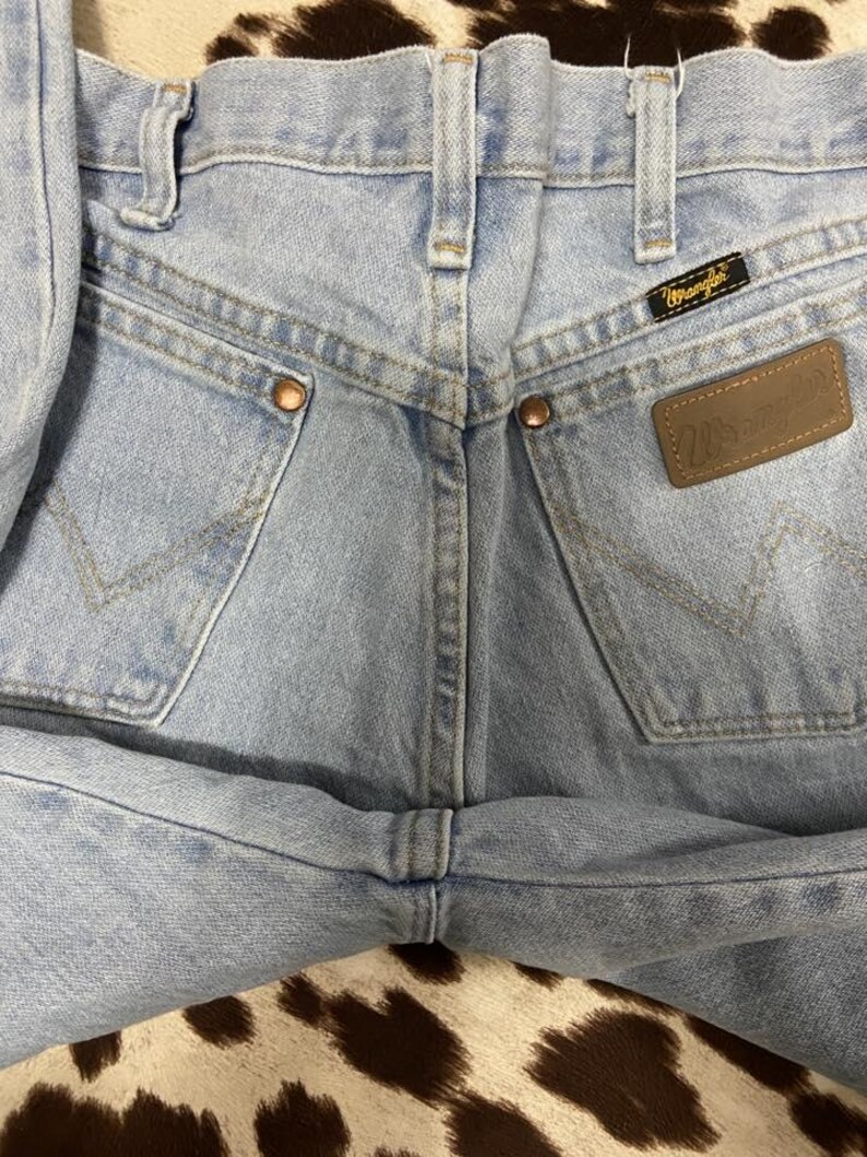 May include: Close-up of light blue denim jeans. The jeans feature classic five-pocket styling, copper rivets, and a brown leather Wrangler patch. The Wrangler label is visible on the back pocket. The jeans are folded.