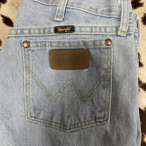 May include: Pale blue denim jeans with a classic five-pocket design. Features a brown leather patch with the "Wrangler" logo on the back pocket. The jeans have copper rivets and a black label with the "Wrangler" logo.