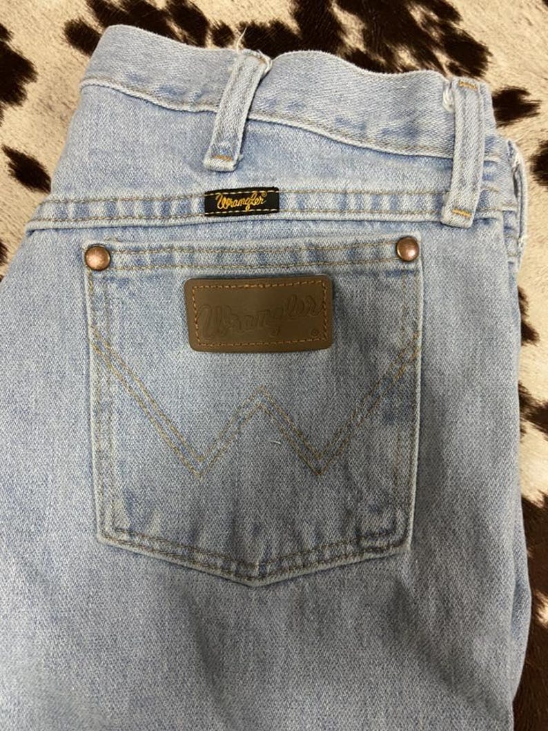 May include: Pale blue denim jeans with a classic five-pocket design. Features a brown leather patch with the "Wrangler" logo on the back pocket. The jeans have copper rivets and a black label with the "Wrangler" logo.
