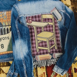 May include: A blue denim jacket featuring a large rectangular woven patch on the back. The patch depicts a yellow school desk with an apple on top against a purple plaid background, with a pencil graphic at the top right and bottom edge.
