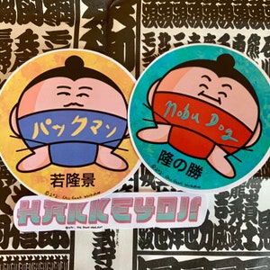 May include: Two round stickers featuring cartoon sumo wrestlers. The first sticker is yellow with a blue and white mask and the text "パックマン" and "若隆景". The second sticker is blue with a red and white mask and the text "Nobu Dog" and "隆の勝". A third sticker with the text "HAKKEYOJI!" is below the other two stickers.