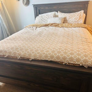 May include: A dark brown wooden bed frame with a matching headboard. The bed is made with a patterned comforter and pillowcases in shades of white and gold. A decorative pillow is also visible.