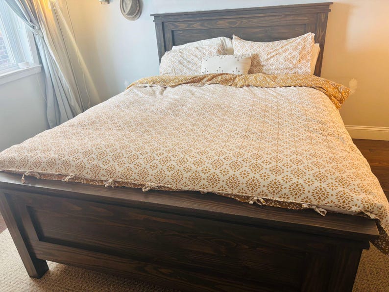 May include: A dark brown wooden bed frame with a matching headboard. The bed is made with a patterned comforter and pillowcases in shades of white and gold. A decorative pillow is also visible.